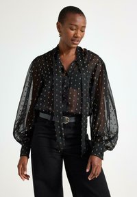 Gennemsigtig sort bluse med guldprikker, lange pufærmer og bundet foran. Båret over sorte bukser med et almindeligt sort bælte.