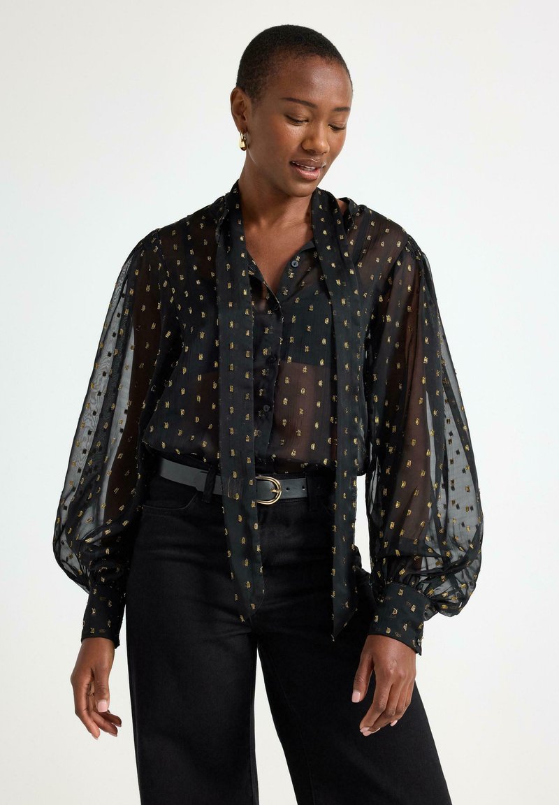 Gennemsigtig sort bluse med guldprikker, lange pufærmer og bundet foran. Båret over sorte bukser med et almindeligt sort bælte.