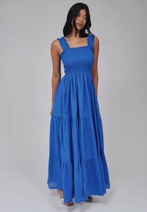 Vrouw met lang zwart haar, gekleed in een mouwloze blauwe maxi-jurk met plooien en beige sandalen, staande tegen een effen grijze achtergrond.