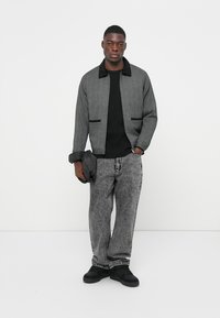 Chaqueta gris con cuello negro, dos bolsillos frontales, llevada sobre un suéter negro texturizado, combinada con unos vaqueros negros deslavados y zapatos negros.
