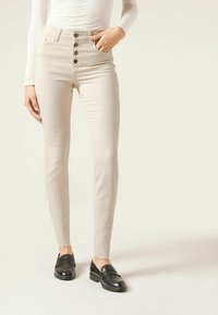 Jeans de tiro alto beige con frente de cinco botones, piernas ajustadas y dobladillo deshilachado, combinados con mocasines negros sobre un fondo plano.