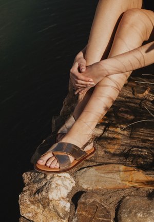 Persoon zittend op rotsachtig oppervlak met gekruiste benen, draagt bruine sandalen, handen geklemd rond één knie, schaduwen vallen op de huid.