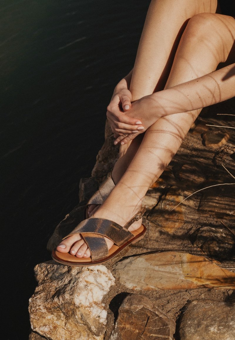 Personne assise sur une surface rocheuse, jambes croisées, portant des sandales marron, mains jointes autour d'un genou, ombres projetées sur la peau.