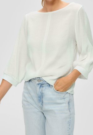 Kobieta ubrana w przezroczystą białą bluzkę z długim rękawem, wsuniętą w jasnoniebieskie, wysokie dżinsy, z jedną ręką w kieszeni.