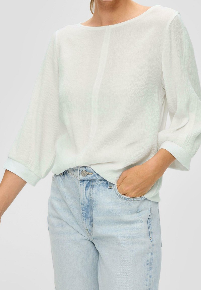 Femme portant un chemisier blanc transparent à manches longues rentré dans un jean en denim bleu clair taille haute, avec une main dans la poche.