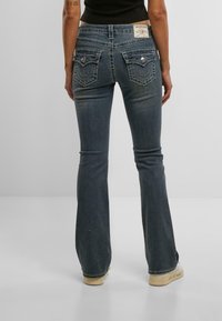 Temno pranje denim flare hlače z našitki, dvema zadnjima žepoma in blagovno znamko na pasu. Teksturirana tkanina, ozek kroj do stegna.