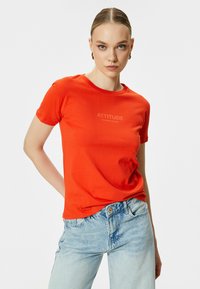 Camiseta roja de manga corta en tela suave con el texto "LA ACTITUD LO ES TODO" en un tono más claro, combinada con unos jeans de color azul claro.