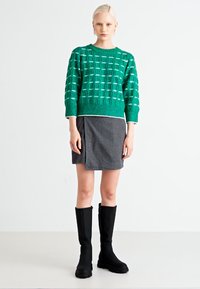 Suéter verde de punto con un patrón de cuadrícula texturizado, combinado con una falda envolvente gris y botas negras hasta la rodilla.