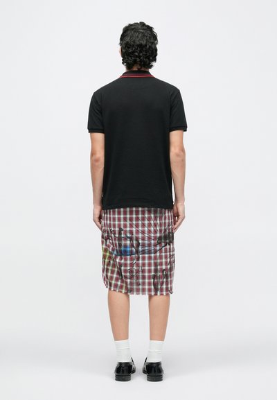 Moški stoji obrnjeno stran, oblečen v črno majico s kratkimi rokavi in rdeče-belo karo krilo do kolen s črnimi abstraktnimi vzorci.