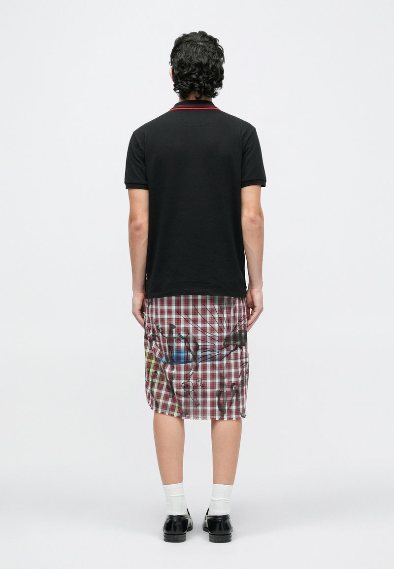 Mand står med ryggen til iført en sort kortærmet skjorte og et knælængde rødt og hvidt ternet skørt med sorte abstrakte mønstre.