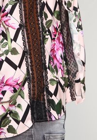 Blusa con stampa floreale caratterizzata da fiori e foglie nei toni del rosa e del verde su uno sfondo rosa chiaro. Dettaglio in pizzo nero e motivo geometrico.