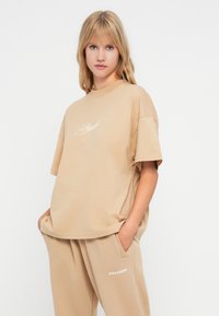 Béžové oversized tričko vyrobené z měkké bavlny, s kulatým výstřihem a krátkými rukávy, na přední straně s vyšitým bílým textem.