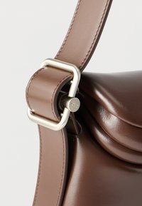 Καφέ δερμάτινος ιμάντας τσάντας με γυαλιστερή ασημένια αγκράφα και καρφίτσα με χαραγμένο "OSSO", προσαρτημένος σε σώμα τσάντας από λείο δέρμα.