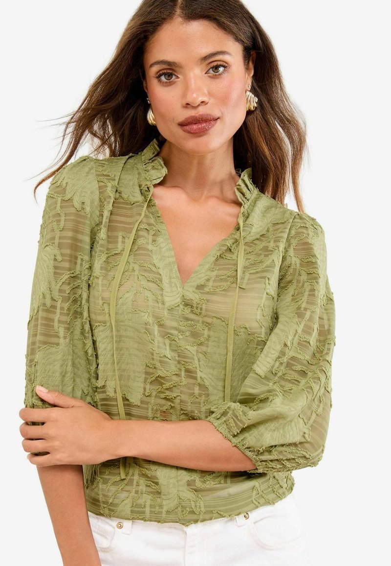 Blusa verde traslucida con tessuto testurizzato, colletto arricciato, dettaglio con cravatta e maniche gather. Design a fantasie con diverse tonalità di verde.