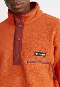 Oranje fleece pullover met een hoge kraag, bruine drukknoopjes, een ritszak op de linkerborst en een Columbia-logo patch.