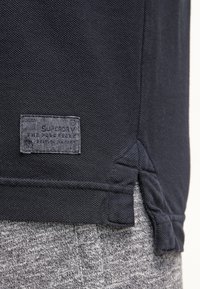 Gros plan sur l'ourlet d'un polo en tissu texturé bleu marine avec une petite étiquette rectangulaire cousue, associé à un pantalon en maille gris clair.