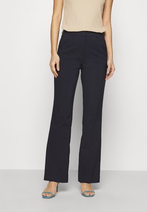 Femme portant un pantalon noir droit, un haut beige sans manches et des sandales à talons hauts ouvertes bleues, debout devant un fond blanc uni.