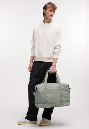 Jonge man in een crème kleurige sweatshirt en zwarte broek, die een lichtgrijze duffeltas met twee handvatten vasthoudt, staande tegen een effen witte achtergrond.