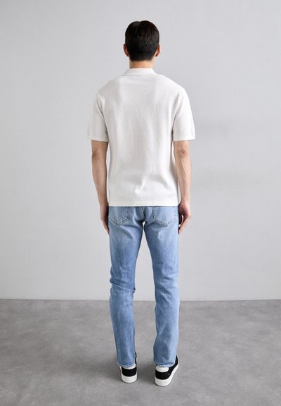 Witte polo met korte mouwen en textuur, gecombineerd met lichtblauwe jeans en zwarte sneakers, gezien vanuit de achterkant tegen een effen achtergrond.