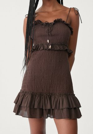 Ærmeløs brun tekstureret kjole med flæsekant, bindebånd og lagdelt kant båret af en person med langt, fletninghår.