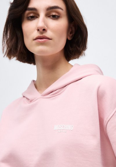 Young woman with short brown hair wearing a light pink Moschino Active hoodie against a plain background.