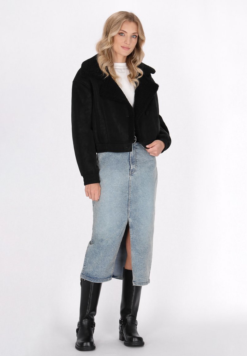 Veste noire courte avec un col en fausse fourrure, associée à une jupe en denim bleu clair avec une fente à l'avant et des bottes noires montantes.