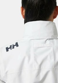 Chaqueta impermeable blanca con un cuello alto, que presenta un logo negro en el hombro y un detalle de bolsillo en la parte trasera. Textura suave.