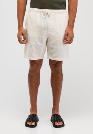Beige bestickte Shorts mit Kordelzugbündchen, versehen mit abstrakten Mustern. Dazu schwarze Riemchensandalen, keine sichtbaren Taschen.