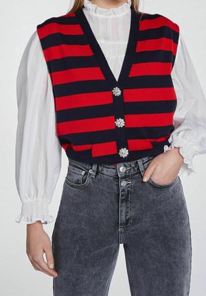 Person iført en rød og sort vandret striped vest med dekorative knapper over en hvid langærmet bluse og høj taljet falmet sorte jeans.