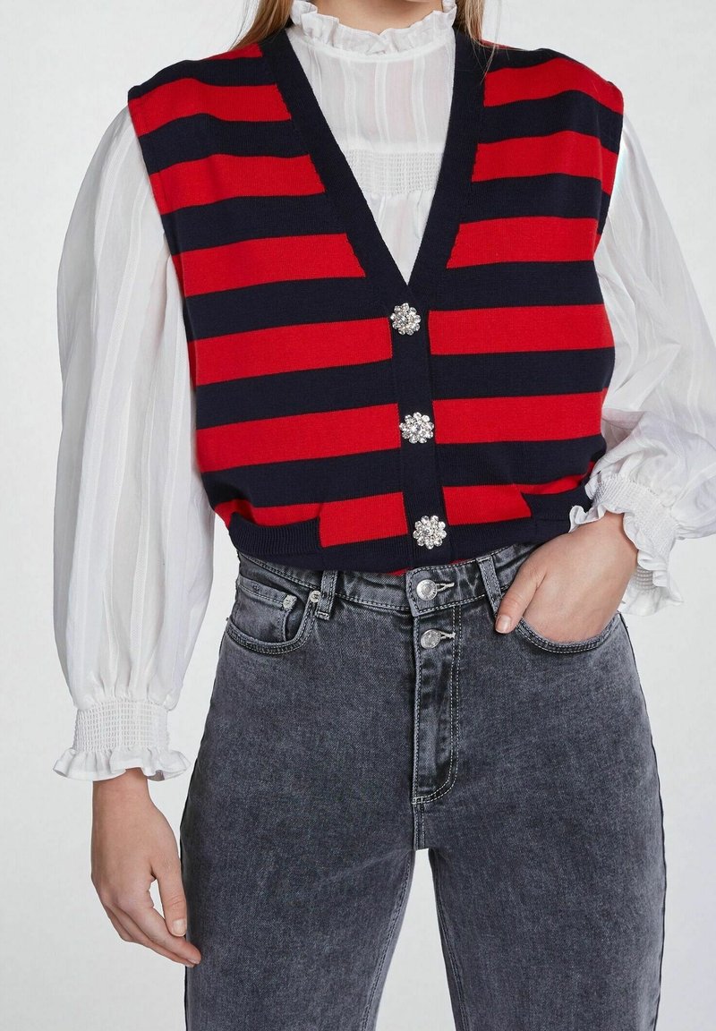Personne portant un gilet rayé horizontal rouge et noir avec des boutons décoratifs sur un chemisier blanc à manches longues et un jean noir délavé taille haute.