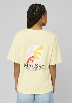 Personne aux cheveux tressés portant un T-shirt jaune pâle avec des formes abstraites colorées et le texte "MATISSE POLYNESIE LE CIEL" au dos.