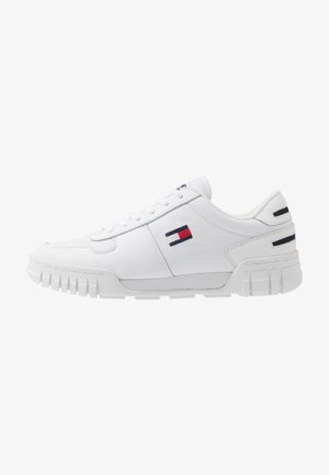 Weißer Sneaker mit niedrigem Schaft, gerillter Sohle und rechteckigem roten, weißen und blauen Logo an der Seite und Ferse, keine sichtbaren gebundenen Schnürsenkel.