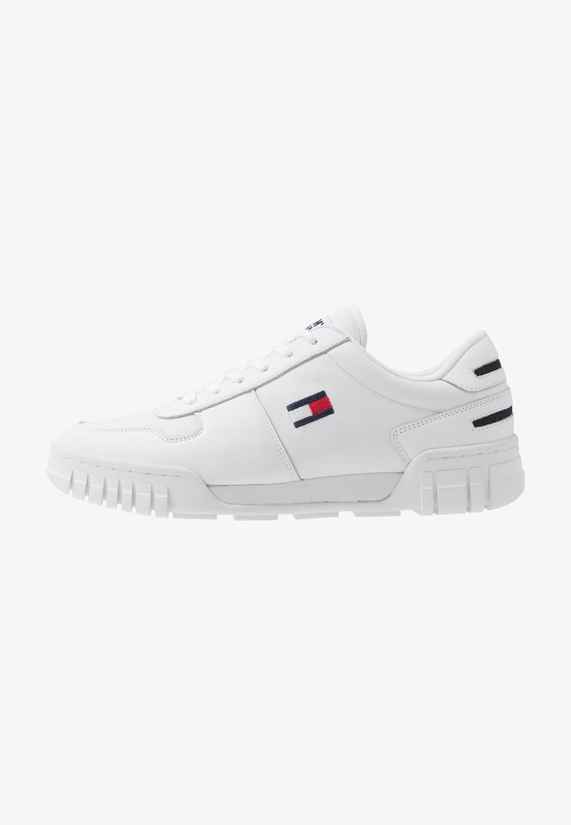 Baskets basses blanches avec semelle rainurée et logo rectangulaire rouge, blanc et bleu sur le côté et le talon, sans lacets visibles noués.