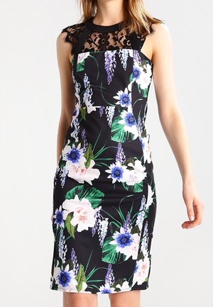 Vrouw draagt een mouwloze zwarte jurk met kanten halslijn en kleurrijke bloemenpatronen in blauw, wit en groen.