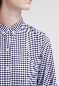 Camicia a quadri blu navy e bianca, colletto a polo, centro bottone corto con bottoni bianchi, tessuto strutturato e dettagli di cucitura ordinati.