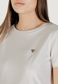 T-shirt beige chiaro in tessuto morbido, con scollo rotondo e una accentatura triangolare in metallo argentato sul petto.