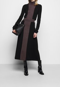 Kvinna klädd i en svart och brun färgblockerad stickad klänning, som håller en svart handväska och bär svarta läder högklackade ankelboots.