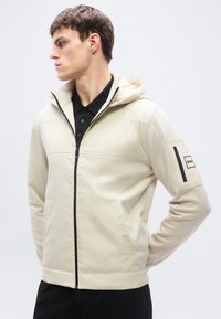 Νεαρός άνδρας που φοράει ανοιχτό μπεζ μπουφάν με κουκούλα, μαύρο φερμουάρ και μπαλώματα με λογότυπο στο αριστερό μανίκι, κοιτάζοντας προς τα αριστερά του.