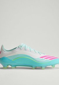 Λευκό και τιρκουάζ ποδοσφαιρικό παπούτσι με ροζ ρίγες, κορδόνια και καλουπωμένες τάπες σε ουδέτερο φόντο.