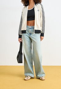 Béžová lehká bunda Adidas na zip s černými pruhy, doplněná světle modrými džíny s vysokým pasem a širokými nohavicemi a béžovými teniskami. Černá taška.