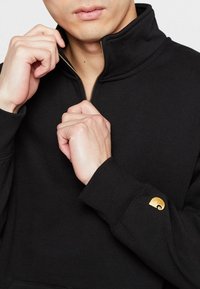 Zwarte zip-up hoodie van zachte stof, met een hoge kraag en gouden logo-accent op de mouw. Slank gesneden met ribgebreide manchetten.