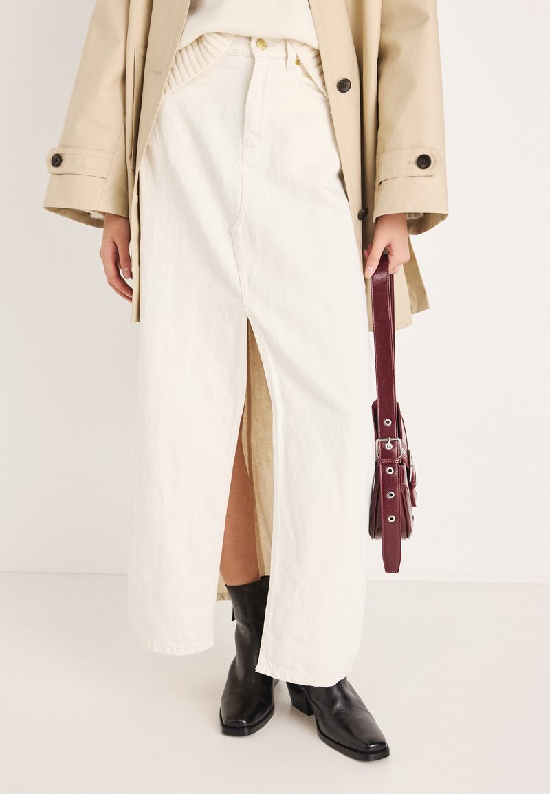 Λευκή μάξι βαμβακερή φούστα με σκίσιμο μπροστά, σε συνδυασμό με μαύρες μποτάκια αστραγάλου. Το μοντέλο κρατάει μια μπορντό τσάντα, φορώντας μπεζ παλτό.