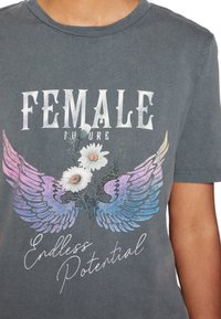 Harmaa lyhythihainen T-paita, jossa on valkoisia ja pastellivärisiä siipigrafiikoita, daisioita sekä tekstit "NAISEN TULEVAISUUS" ja "LOPPUTTOMAT MAHDOLLISUUDAT."