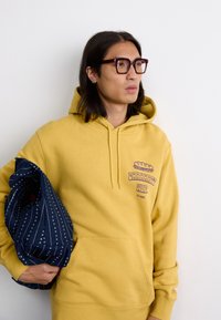 Gelbe Kapuzenjacke mit Fronttasche und Carhartt WIP-Logo. Das Model hält eine marineblaue Tasche aus Stoff mit weißem Muster, vor einem weißen Hintergrund.