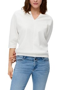 Weiße, strukturierte Bluse mit V-Ausschnitt und Dreiviertelärmeln, kombiniert mit hellblauen Jeans. Die Bluse hat ein dezentes Muster.