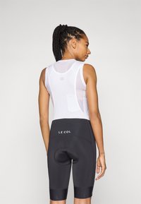 Weißes ärmelloses Fahrradtrikot aus Netzstoff mit Rückentasche, kombiniert mit schwarzen gepolsterten Shorts mit einem Logo an der Taille und den Beinabschlüssen.