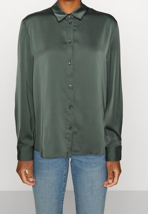 Femme portant une chemise boutonnée en satin vert foncé à manches longues et un jean bleu clair, debout devant un fond clair uni.