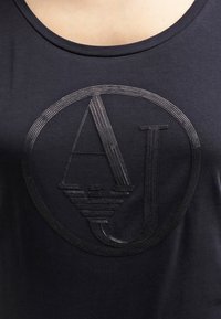 Musta puuvillainen tanktoppi, jossa on teksturoitu musta "AJ" -logo ympyrämuotoisessa designissa, pyöreä pääntie ja hihattomat lähestymistavat.