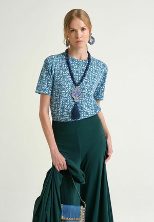 Donna che indossa una maglia blu con motivo, gonna verde scuro, tiene una giacca abbinata e una borsa blu, accessoriata con collana e orecchini blu.