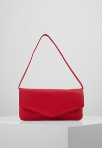 Červená texturovaná kožená kabelka ve stylu obálky s tenkým popruhem vystavená na bílém podstavci na šedém pozadí.